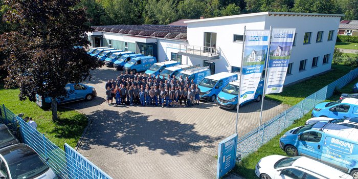 Eine große Gruppe von Mitarbeitern steht vor einem modernen Gebäude, umgeben von blauen Lieferwagen und grüner Landschaft.