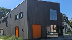 Ein modernes Gebäude mit dunkler Holzverkleidung und großen Fenstern, umgeben von grünen Bäumen und einem blauen Himmel.