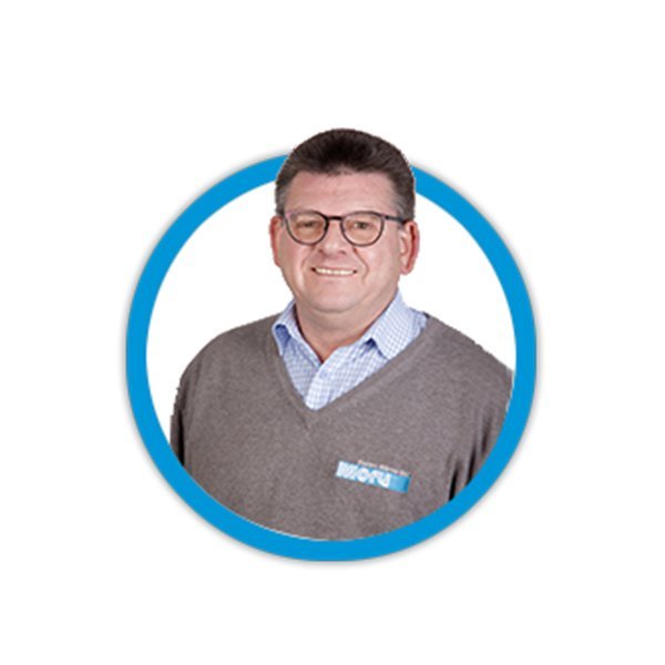 Ein lächelnder Mann mit Brille trägt einen grauen Pullover und ein kariertes Hemd, umrahmt von einem blauen Kreis.