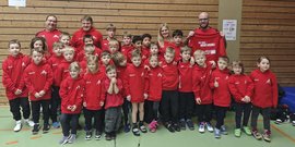 Eine Gruppe von Kindern in roten Hoodies posiert fröhlich in einer Sporthalle, umgeben von Holzverkleidung und Sportgeräten.