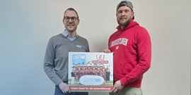 Zwei Männer stehen vor einer hellen Wand und halten ein großes, signiertes Teamfoto mit Dankesbotschaft in den Händen.