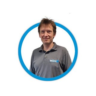 Ein Mann mit braunen Haaren lächelt freundlich in einem grauen Poloshirt mit einem Logo auf der linken Brustseite.