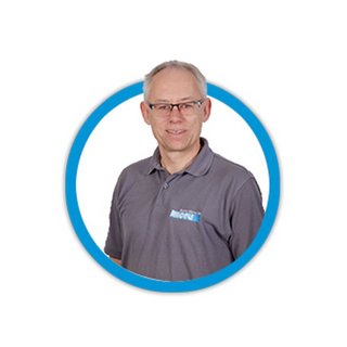 Ein lächelnder Mann mit Brille trägt ein graues Polohemd und steht vor einem weißen Hintergrund in einem runden Rahmen.