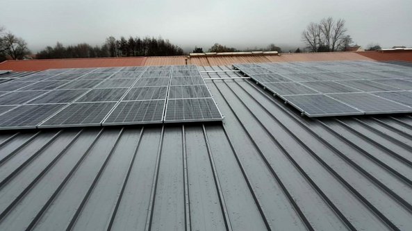 Auf einem grauen Metalldach sind mehrere Solarpanels installiert, während der Himmel bewölkt und trüb ist.
