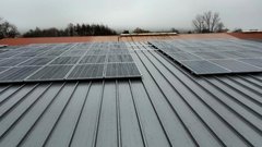 Auf einem grauen Metalldach sind mehrere Solarpanels installiert, während der Himmel bewölkt und trüb ist.