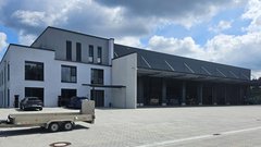 Ein modernes Gebäude mit großen Fenstern und einem überdachten Bereich, umgeben von einem gepflasterten Parkplatz.
