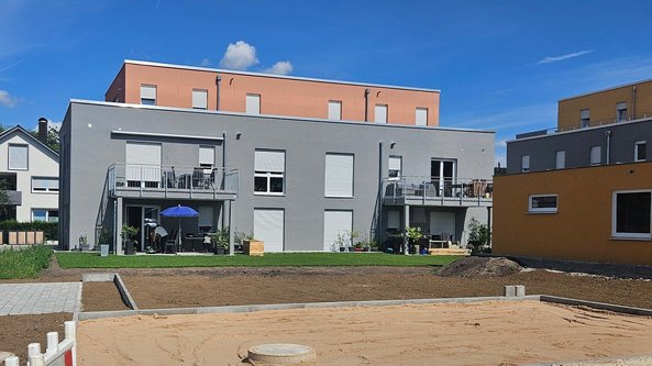 Zwei moderne Wohngebäude mit Balkonen und einem blauen Sonnenschirm stehen in einem neu gestalteten Gartenbereich.