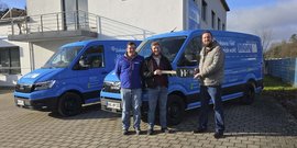 Drei Männer stehen vor zwei blauen Lieferwagen, einer hält einen Schlüssel in der Hand, während sie lächeln.