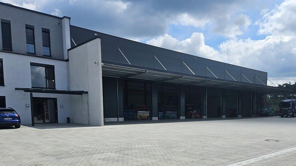 Ein modernes Lagergebäude mit großen Fenstern und einem überdachten Eingangsbereich, umgeben von einem gepflasterten Parkplatz.