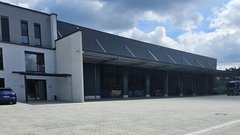 Ein modernes Lagergebäude mit großen Fenstern und einem überdachten Eingangsbereich, umgeben von einem gepflasterten Parkplatz.