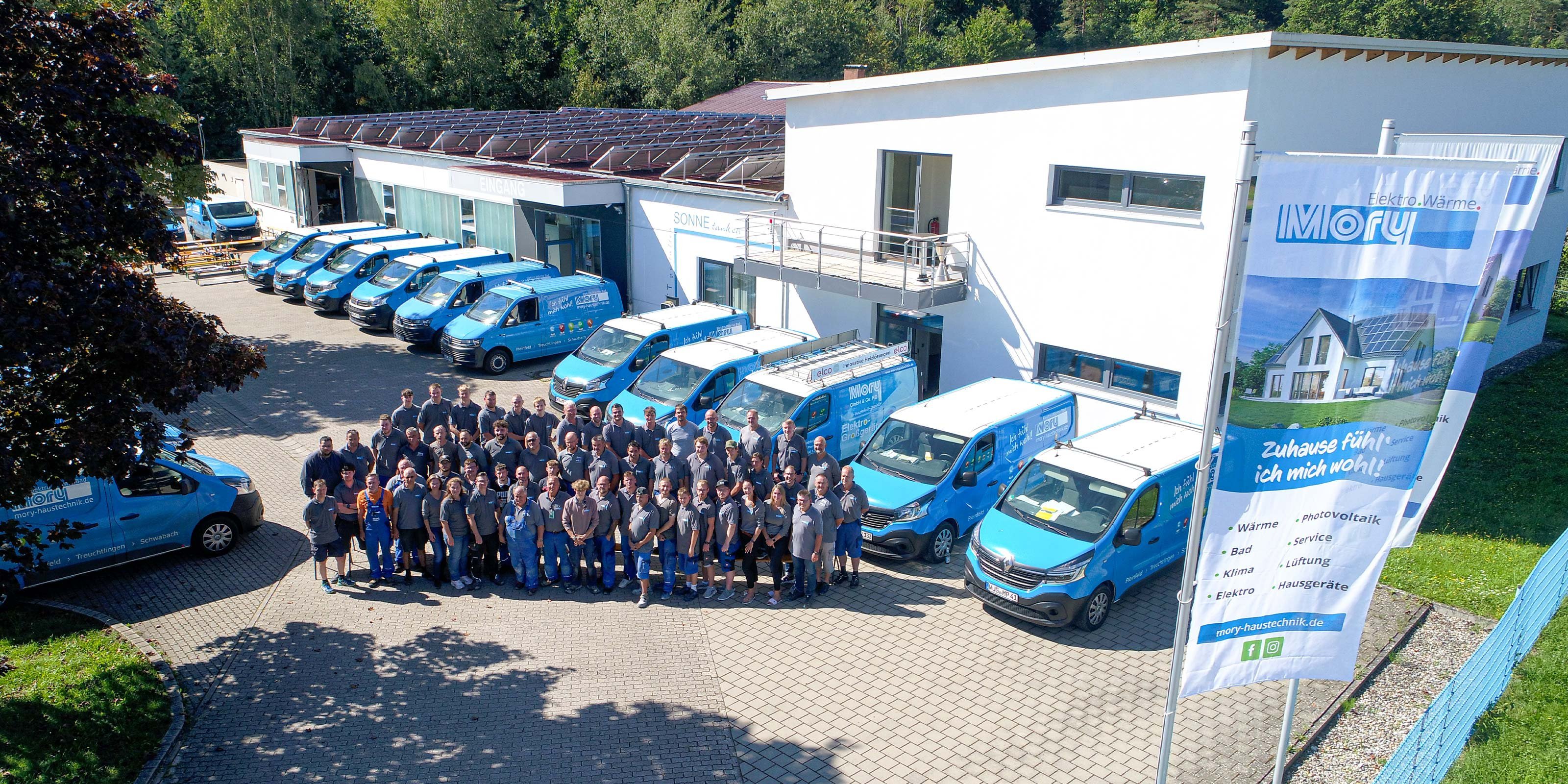 Das Team von Moray Haus Technik posiert lächelnd vor ihrem Firmengebäude, während mehrere blaue Fahrzeuge im Hintergrund parken.
