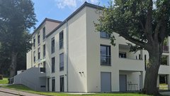 Ein modernes, helles Gebäude mit großen Fenstern und einem begrünten Außenbereich, umgeben von Bäumen und einer ruhigen Straße.
