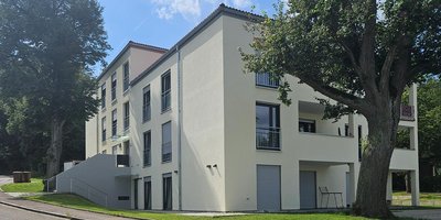 Ein modernes, helles Gebäude mit großen Fenstern und einem begrünten Außenbereich, umgeben von Bäumen und einer ruhigen Straße.