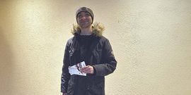 Ein junger Mann mit einem Lächeln steht vor einer hellen Wand und hält mehrere Tickets in der Hand.
