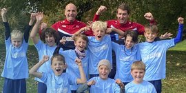 Eine Gruppe von Kindern in blauen Trikots jubelt fröhlich, während zwei Trainer im Hintergrund stolz lächeln.