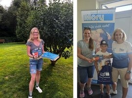 Im Vordergrund lächelt eine Frau mit langen Haaren, die ein Plakat und einen Preis in der Hand hält, während sie von zwei anderen Personen umgeben ist.