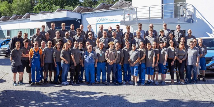 Eine große Gruppe von Mitarbeitern steht lächelnd vor einem Gebäude mit dem Schriftzug 'EINGANG' und 'SONNE tanken'.