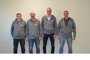 Vier Männer stehen nebeneinander vor einer hellen Wand, alle tragen graue Kapuzenpullover und lächeln freundlich.