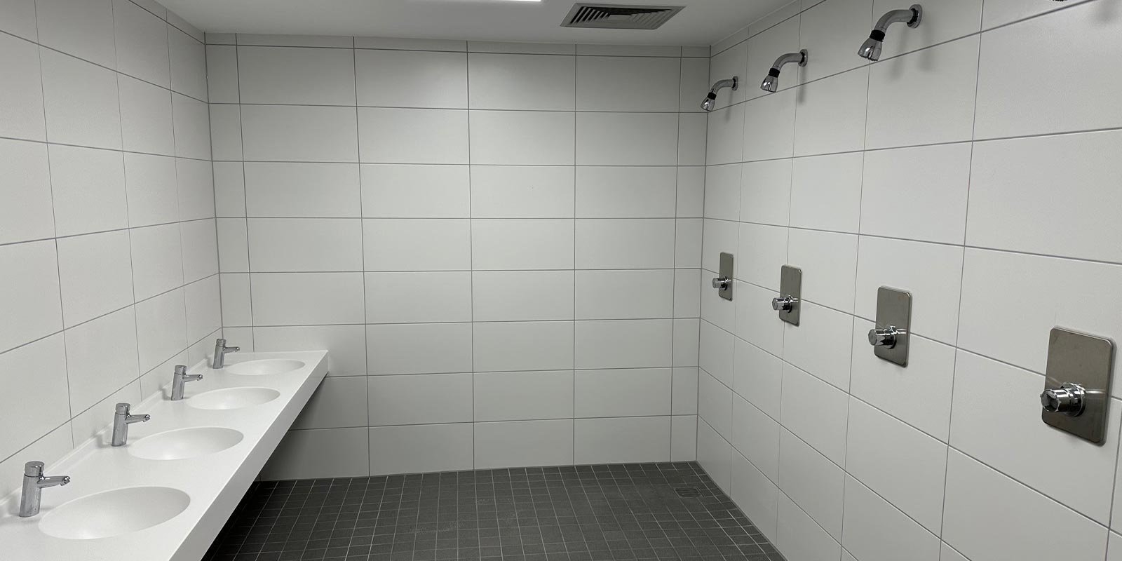 Ein modernes Badezimmer mit weißen Fliesen, das fünf Waschbecken und mehrere Duschköpfe an der Wand zeigt.