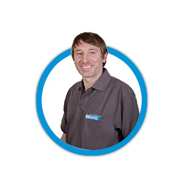 Ein lächelnder Mann trägt ein graues Polohemd mit einem blauen Logo, umgeben von einem blauen Kreis.