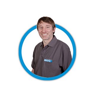 Ein lächelnder Mann trägt ein graues Polohemd mit einem blauen Logo, umgeben von einem blauen Kreis.