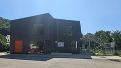 Ein modernes, zweigeschossiges Gebäude mit Holzverkleidung und großen Fenstern, umgeben von Bäumen und einem blauen Himmel.