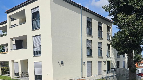 Die Fassade eines hellen Wohnhauses mit Rollläden und einem einladenden Eingangsbereich, der von einem Baum beschattet wird.