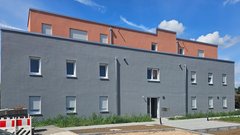 Ein modernes Gebäude mit grauer Fassade und einem oberen Stockwerk in sanftem Orange, umgeben von grünem Gras und einem blauen Himmel.