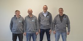 Vier Männer stehen nebeneinander vor einer hellen Wand, alle tragen graue Kapuzenpullover mit einem Logo.
