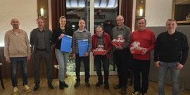 Die Gruppe lächelt fröhlich, während sie Geschenke und Dokumente in einem gemütlichen, weihnachtlich geschmückten Raum präsentiert.
