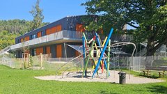 Ein moderner Spielplatz mit bunten Klettergerüsten steht vor einem hellen, zweigeschossigen Gebäude in einer grünen Umgebung.