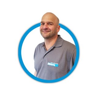 Ein lächelnder Mann trägt ein graues Poloshirt mit einem blauen Logo, umgeben von einem hellen, runden Hintergrund.