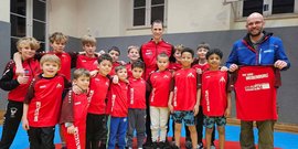 Eine Gruppe von Jungen in roten Sporttrikots posiert fröhlich in einer Sporthalle, während ein Trainer ein Trikot hält.