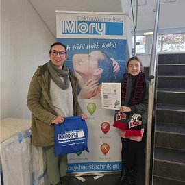 Zwei Frauen stehen vor einem Werbebanner, eine hält eine Broschüre und die andere eine Tasche in der Hand.