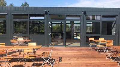 Die Terrasse eines stilvollen Cafés, umgeben von Bäumen, mit mehreren Holzstühlen und einem klaren blauen Himmel im Hintergrund.