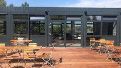 Die Terrasse eines stilvollen Cafés, umgeben von Bäumen, mit mehreren Holzstühlen und einem klaren blauen Himmel im Hintergrund.