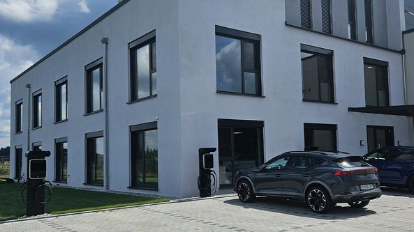 Ein modernes Gebäude mit großen Fenstern und einem gepflegten Rasen, auf dem zwei Elektroautos parken.
