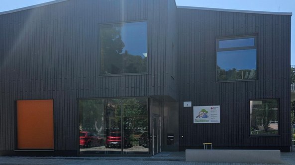 Ein modernes, zweigeschossiges Gebäude mit dunkler Holzverkleidung und großen Fenstern, umgeben von grüner Natur.
