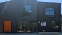 Ein modernes, zweigeschossiges Gebäude mit dunkler Holzverkleidung und großen Fenstern, umgeben von grüner Natur.