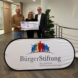 Drei Personen stehen in einem modernen Raum und halten einen großen Scheck über 100.000 Euro für die BürgerStiftung.
