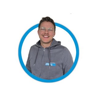 Der Mann mit einem freundlichen Lächeln hat kurze Haare und trägt eine graue Jacke mit einem blauen Logo auf der Brust.