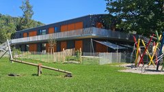Ein modernes Gebäude mit orangefarbenen Fenstern steht in einem grünen Garten, umgeben von einem Zaun und Spielgeräten.